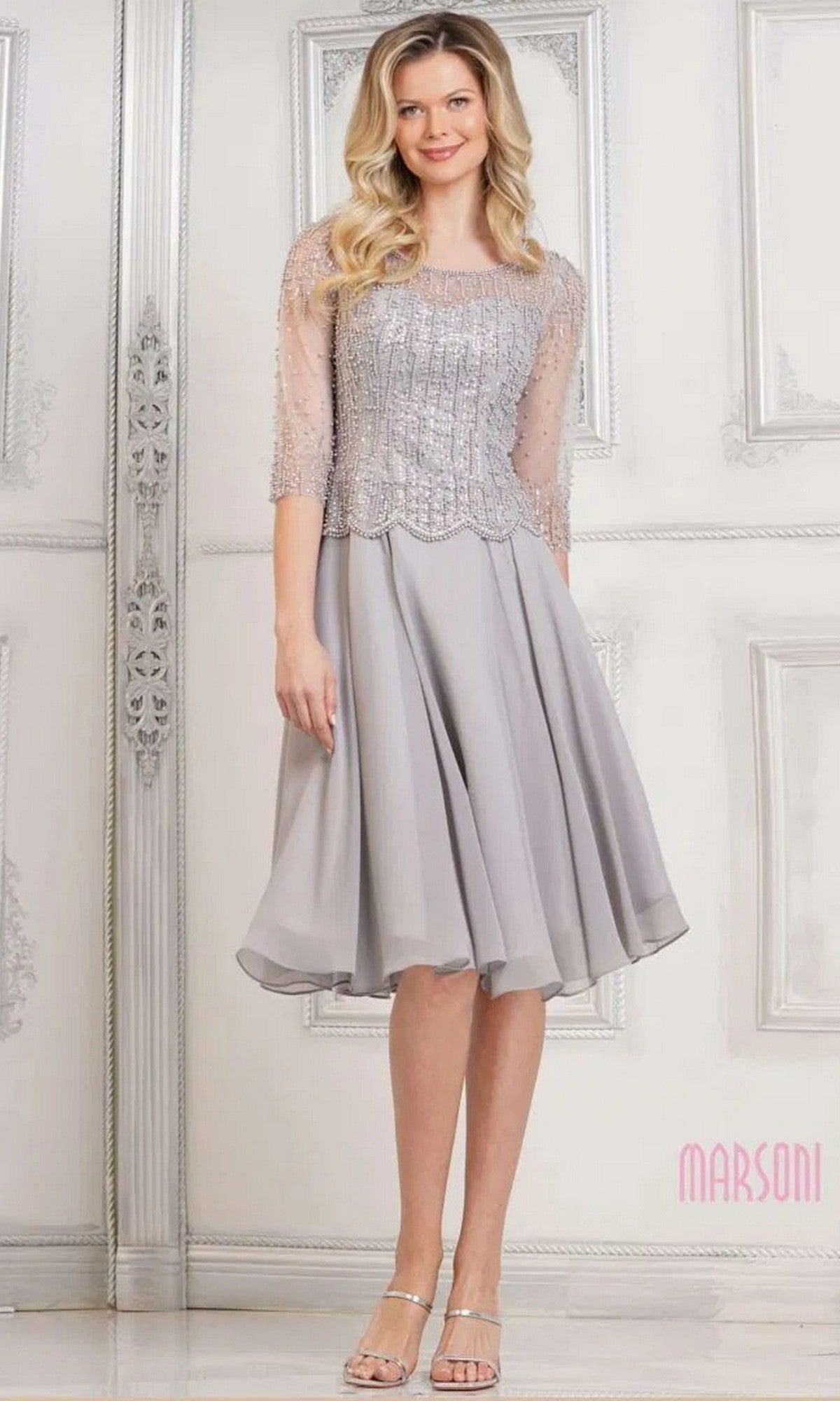 A woman wears the Marsoni M312S knee-length semi-formal dress in grey, featuring an embellished bodice, sheer sleeves, and a flowy skirt, paired with silver sandals.